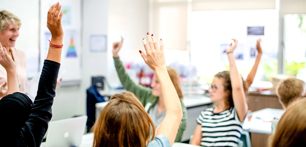 Students raising hands eagerly in a bright classroom with smiling teacher