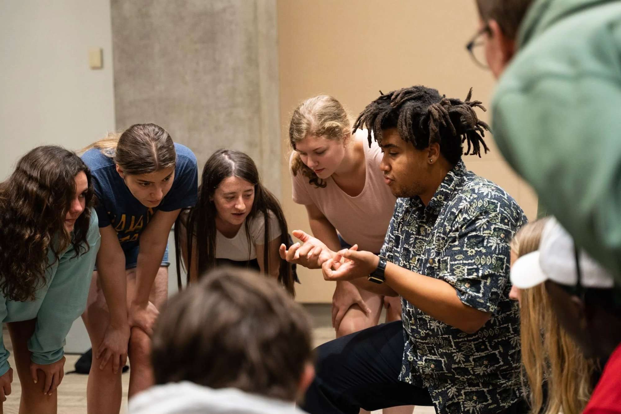 A group of young adults leaning in attentively to listen to a young man speaking and gesturing with his hands.
