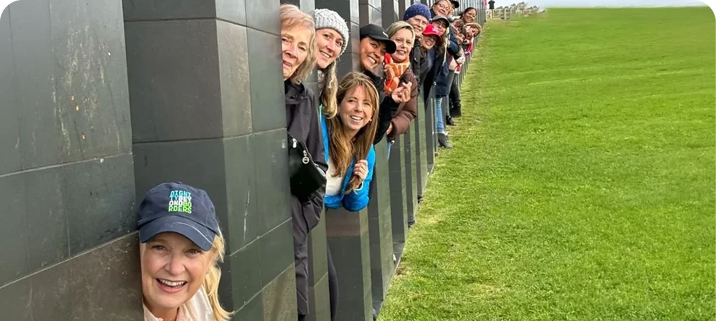 Group of smiling women peeking around a wall outdoors on a green lawn