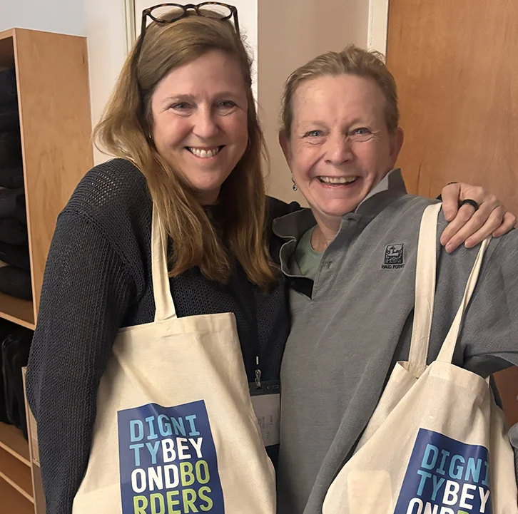 Two smiling women holding Dignity Beyond Borders canvas tote bags at a community gathering.