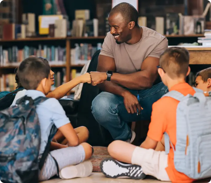 Teacher fist-bumping a student while sitting with a group of children in a school library.