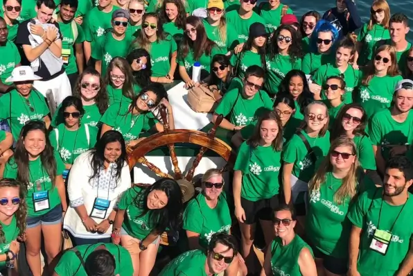 Large group of students in green Common Ground T-shirts posing around a ship's wheel outdoors