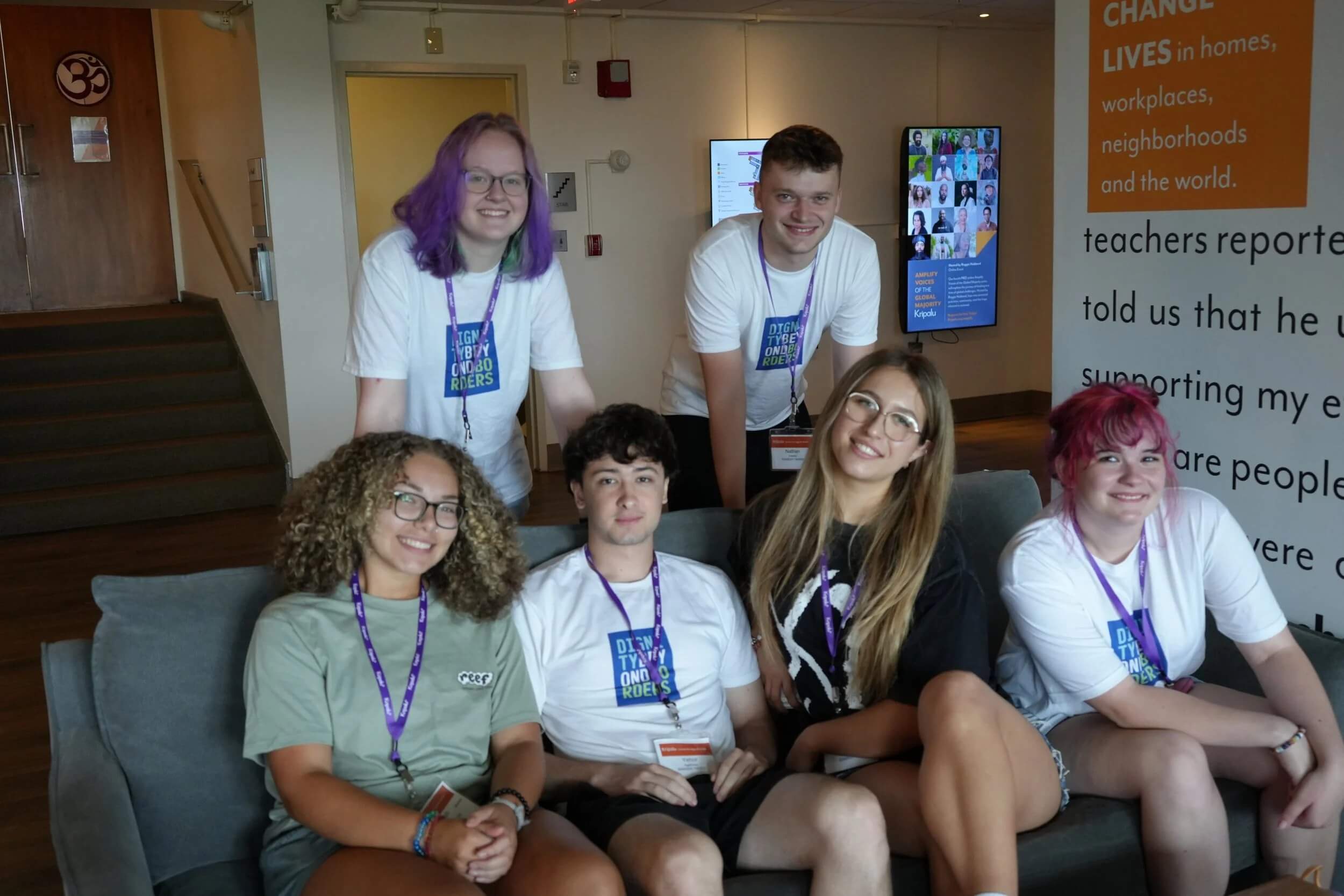 A group of six smiling young adults posing together on a couch indoors.