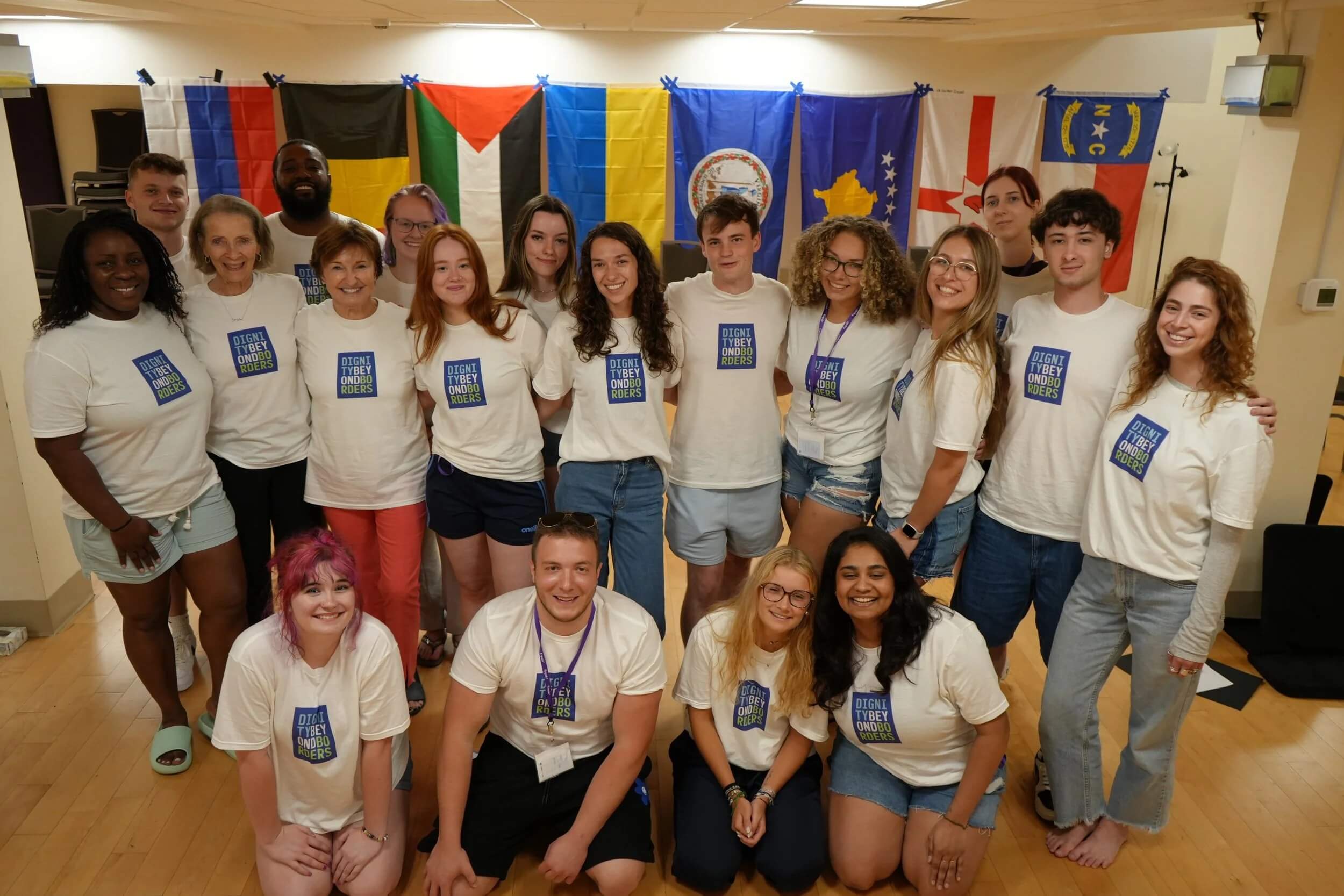 A large, diverse group of participants standing together in matching t-shirts in front of a wall of international flags.