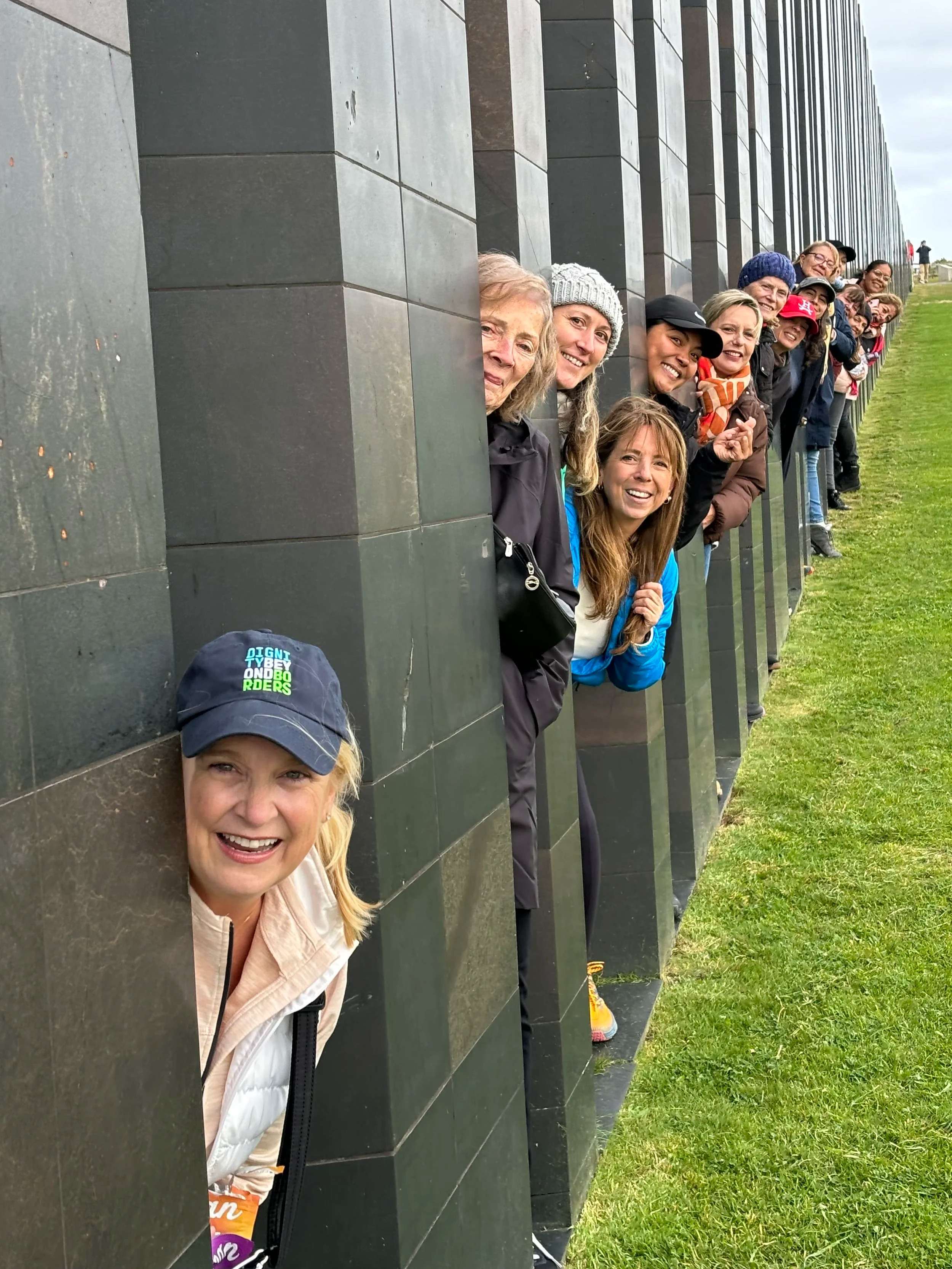 A long line of smiling women playfully peeking out from behind a row of large stone pillars, with the woman in front wearing a Dignity Beyond Borders cap.