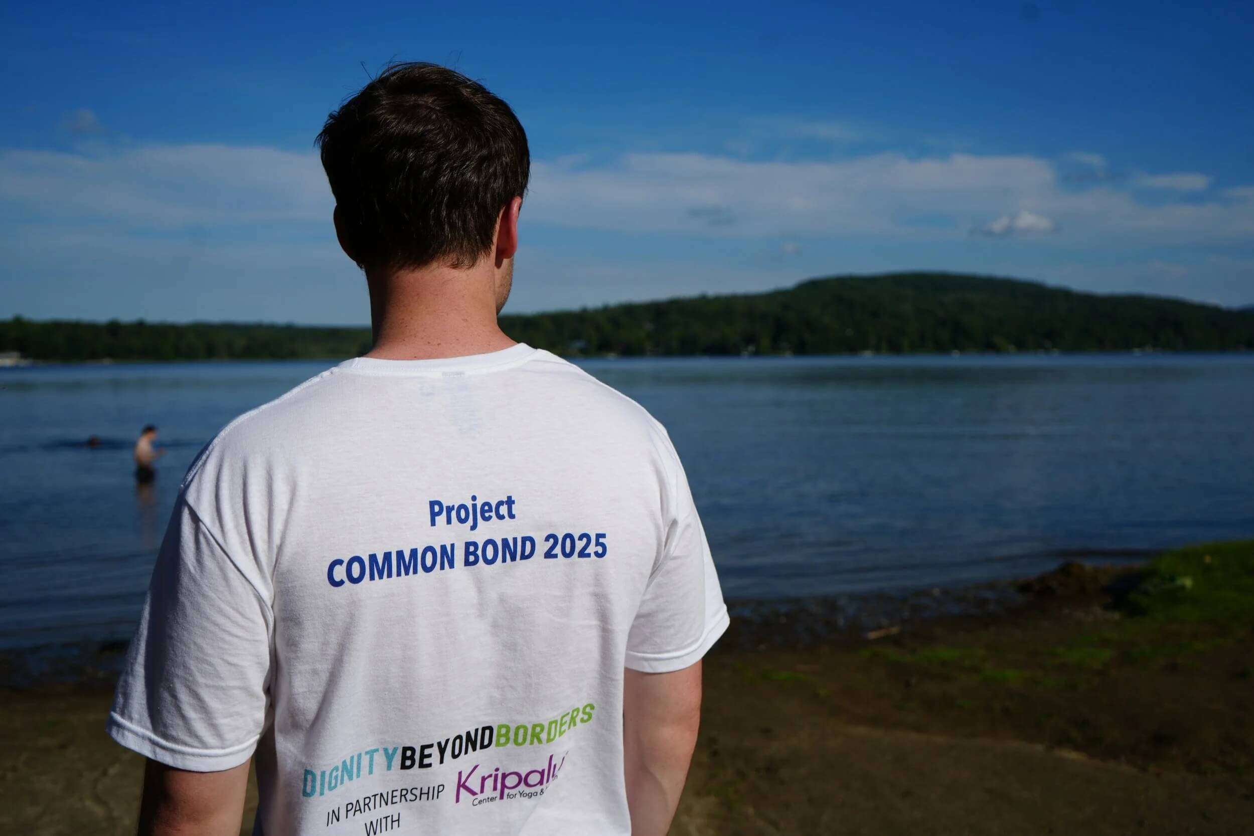 A participant seen from behind wearing a Project Common Bond t-shirt, looking out over a calm, scenic lake.