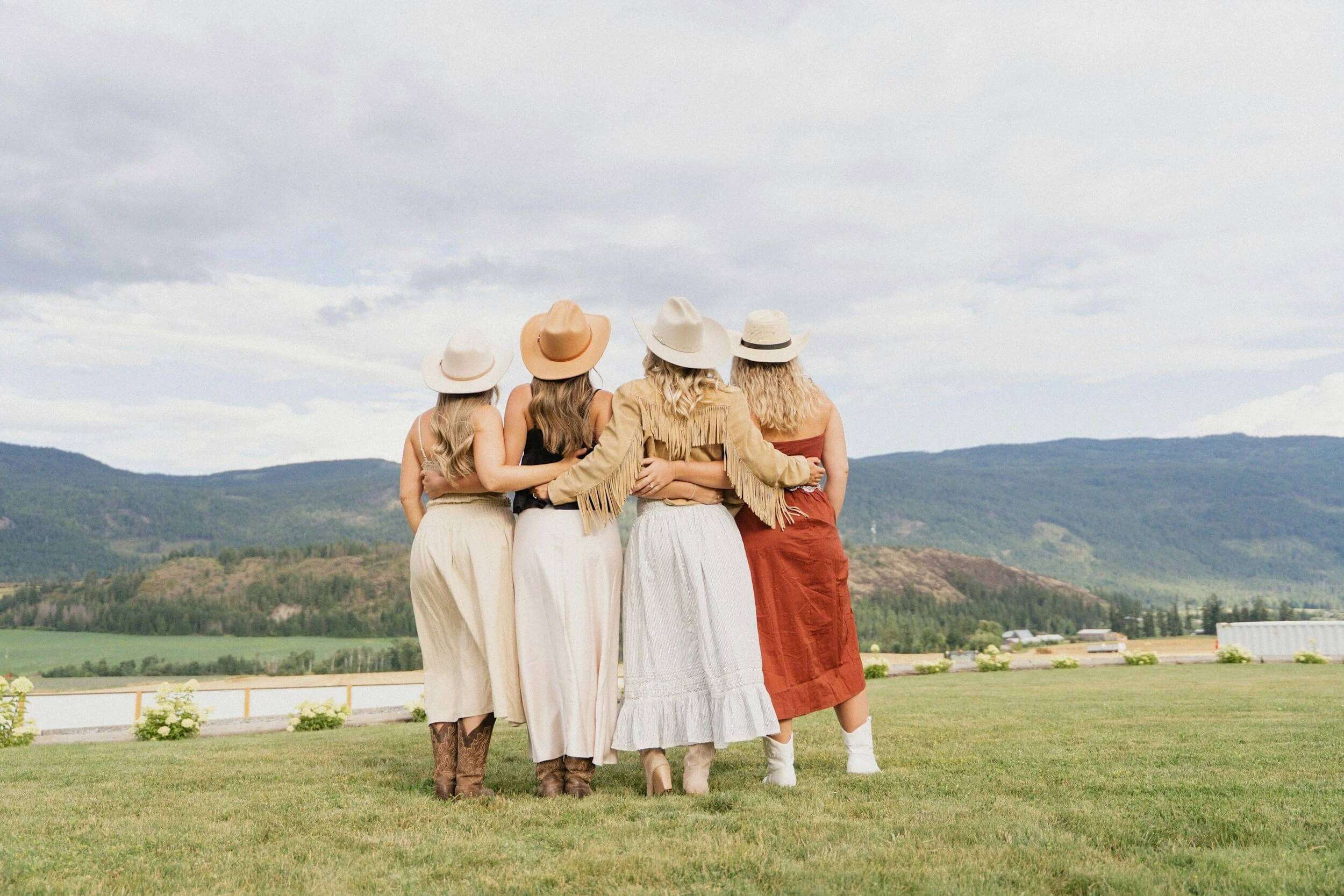 Four women in cowboy hats standing arm-in-arm, looking out over a wide mountain landscape.