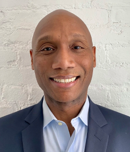 Michael Frazier smiling in a navy blazer against a white brick wall