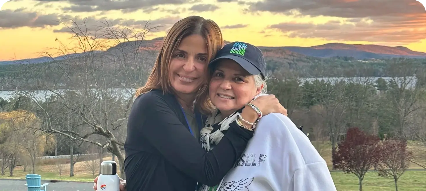 Two smiling women hugging warmly outdoors, with one wearing a Dignity Beyond Borders baseball cap.