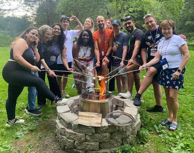 Group of people roasting marshmallows around a campfire A diverse group of smiling people stand in a circle outdoors, roasting marshmallows on long sticks over a metal fire pit.