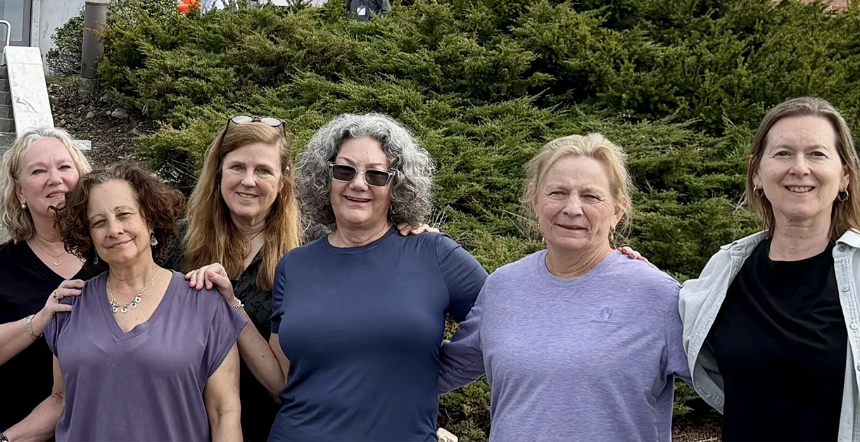 Six women smiling together outdoors in front of evergreen shrubs