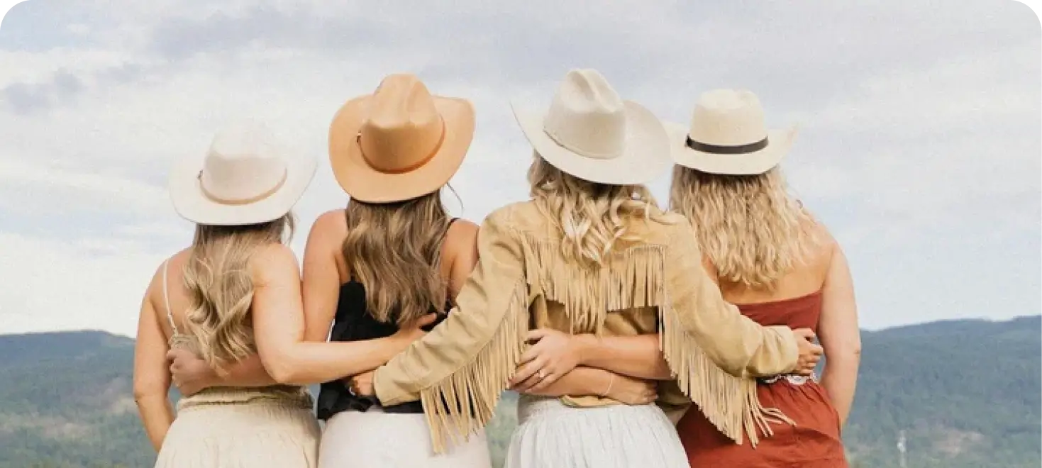 Four women in cowboy hats standing arm-in-arm, looking out over a beautiful mountain landscape.
